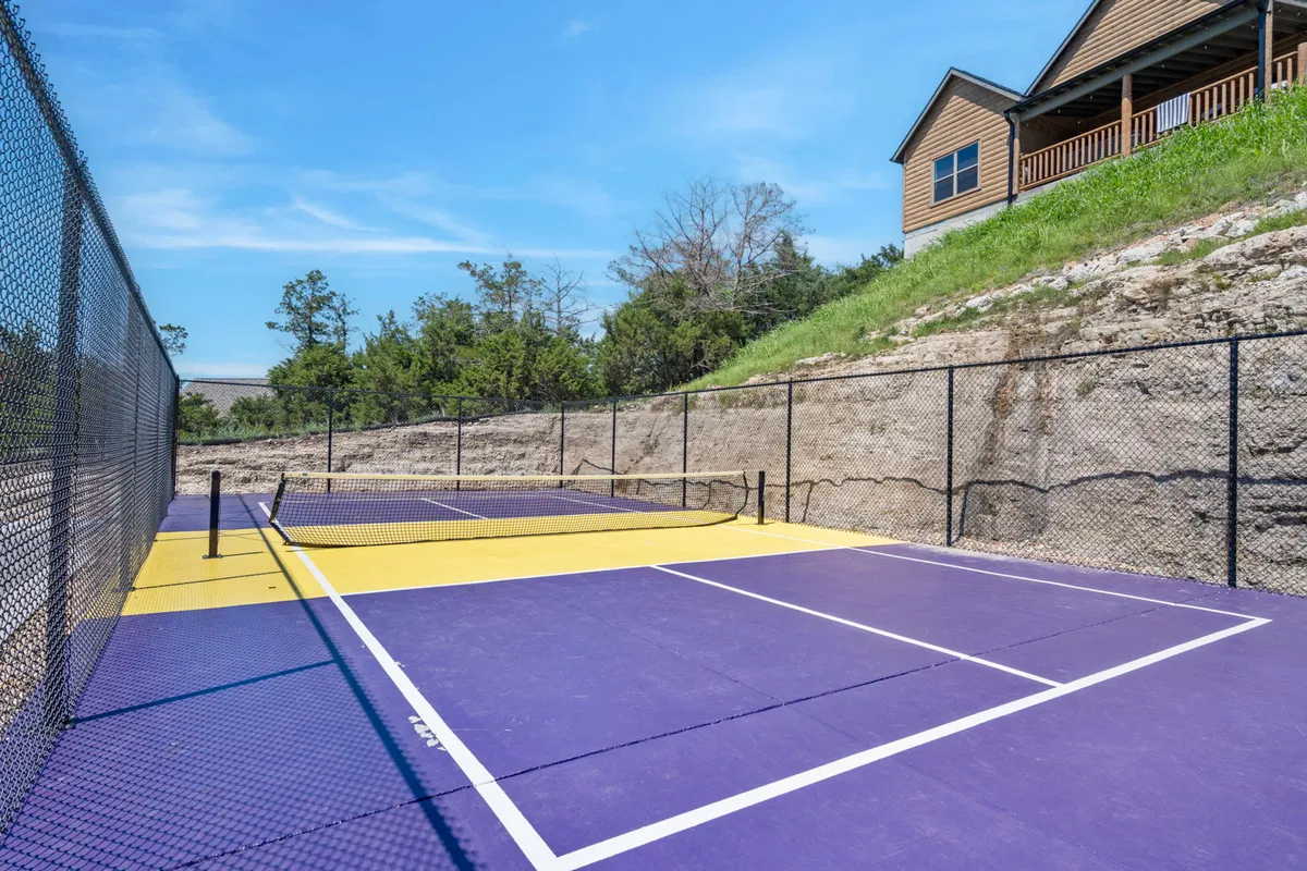 This image features a vibrant tennis court surrounded by lush greenery with a cabin visible in the background. The recreational setting is lively and promotes outdoor activities like pickleball or tennis, complementing the active adventure theme of the section.