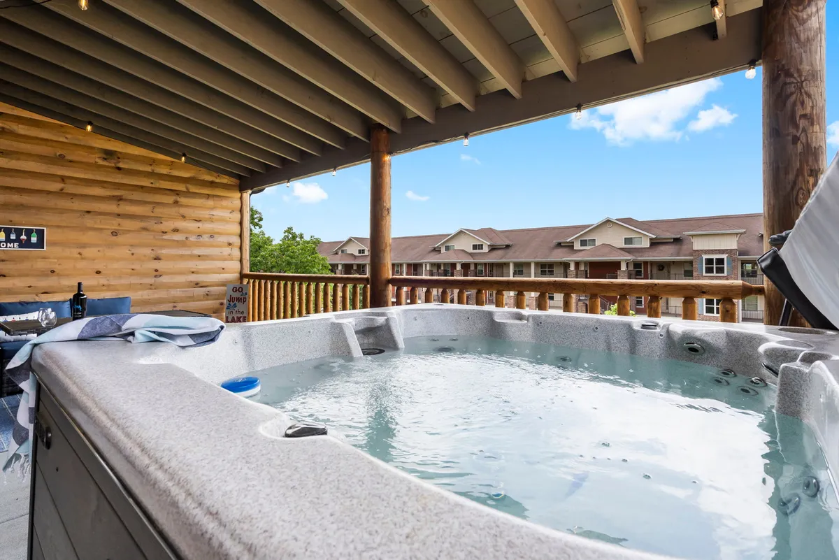 An inviting hot tub sits on an enclosed wooden deck, surrounded by a sturdy railing and set against a backdrop of scenic natural views and a wooden cabin wall. The image conveys a sense of relaxation and privacy, with space for gathering or unwinding in comfort. Visible hints of activity include people enjoying the hot tub and appreciating the view, suggesting both social and peaceful moments.