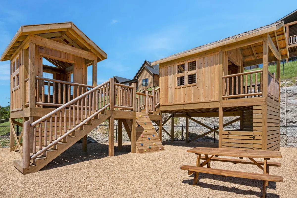 A playful, colorful outdoor play structure with a climbing wall and a wooden picnic table, set in a sunlit, grassy area. The scene exudes energy, joy, and the promise of imaginative, active play for children.