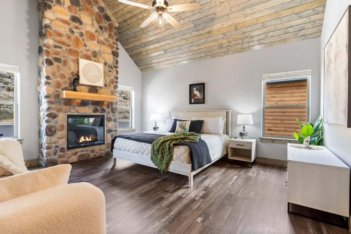 A tranquil bedroom featuring a large bed positioned near a stone fireplace, with a ceiling fan overhead and expansive windows letting in natural light. The space exudes warmth and relaxation, inviting guests to unwind in style.