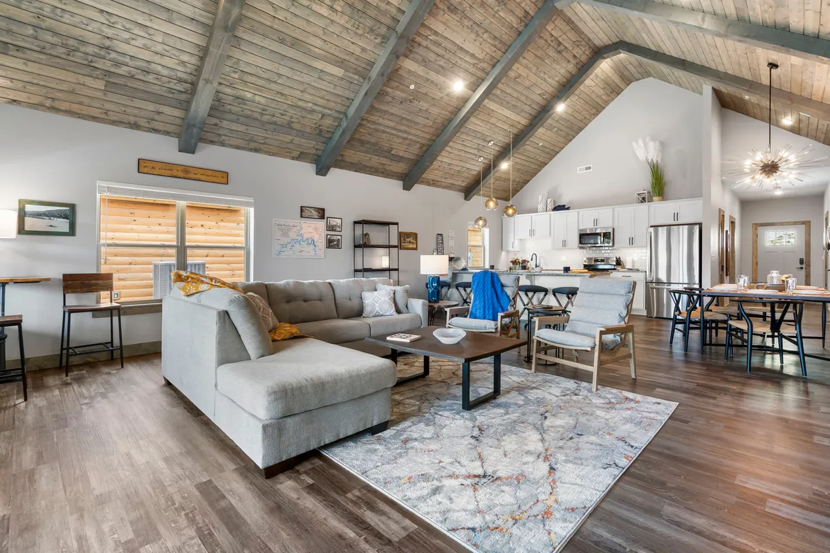 A wide-angle interior view connecting the comfortable sectional sofa, a modern coffee table, a decorative rug, a bright dining area with chairs, and a kitchen island in the background. The layout emphasizes an open, seamless flow between living, dining, and kitchen areas.