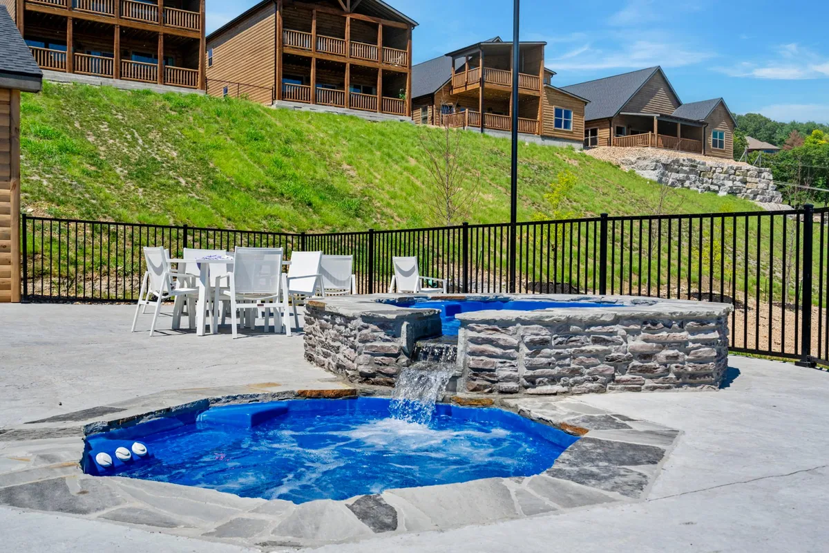 An exterior view featuring a private hot tub surrounded by a tranquil setting, a waterfall feature, outdoor dining table, and neighboring cabins. The scene captures the essence of both relaxation and outdoor socialization, perfect for unwinding or gathering with friends and family under sunset skies.