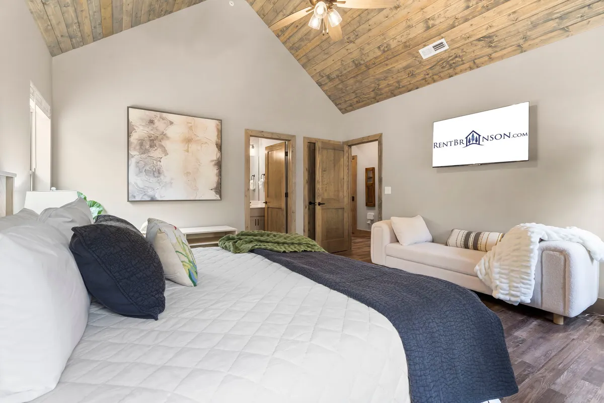 A serene bedroom showcasing a spacious bed with decorative pillows and a textured throw blanket. Artwork adorns the walls, and a TV is mounted opposite the bed. The room is filled with soft, natural light, creating a calming atmosphere.