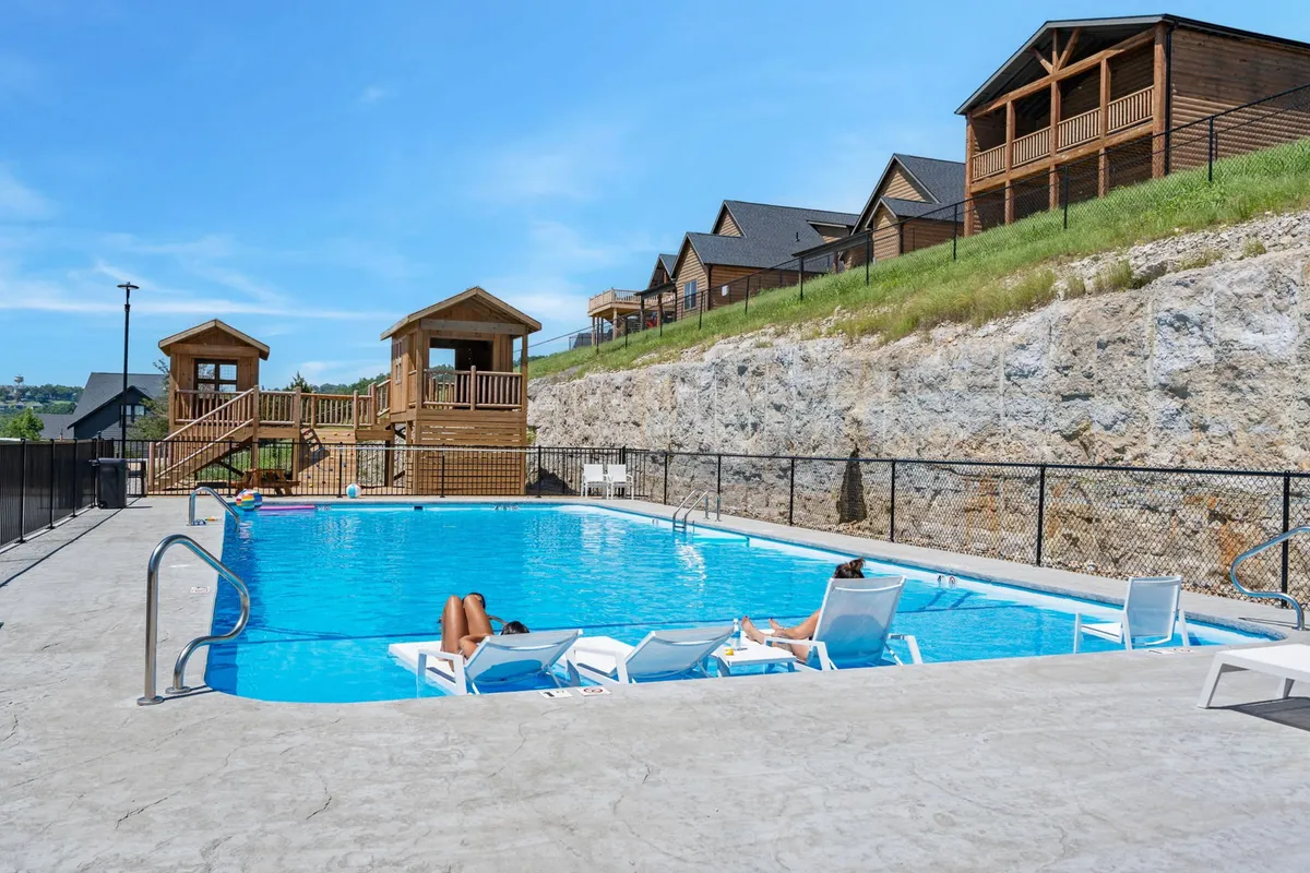 An outdoor scene with a sparkling swimming pool, poolside loungers, and a cheerful playground structure in the background nestled among cabins. The setting feels lively and social, with families and children enjoying sunny days, playtime, and refreshing swims.