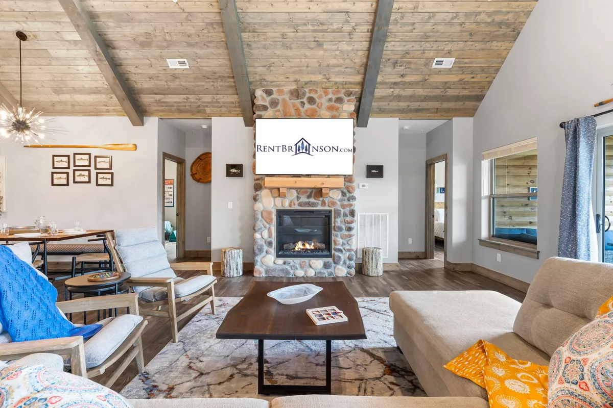 This image showcases the warm and inviting interior of a log cabin, featuring a crackling stone fireplace, plush seating arranged for gatherings, a spacious dining table perfect for shared meals, and elegant rustic décor that celebrates Ozark charm. The cozy ambiance suggests comfort, relaxation, and togetherness.