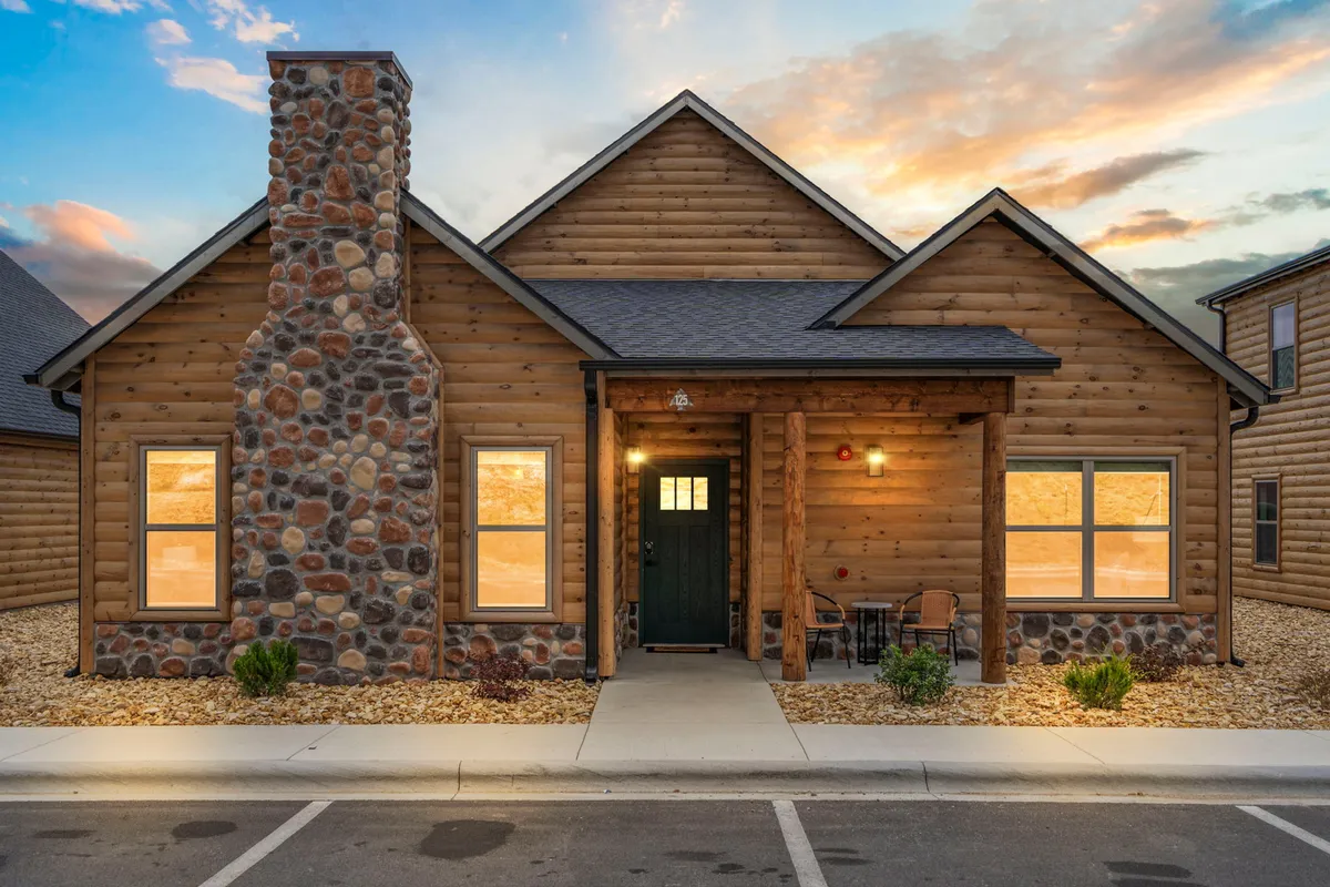 A cozy exterior view of the home featuring a stone chimney, wooden facade, and an inviting porch with seating. The scene evokes a sense of arrival, warmth, and togetherness—perfect for family gatherings, evening chats, or simply watching the sun set over the surrounding landscape.